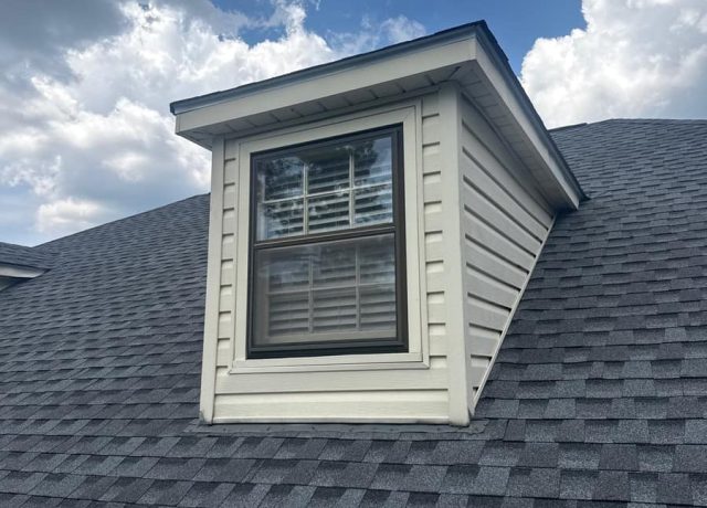 Cream-sided dormer with a window, shingles running to its base under blue sky and clouds